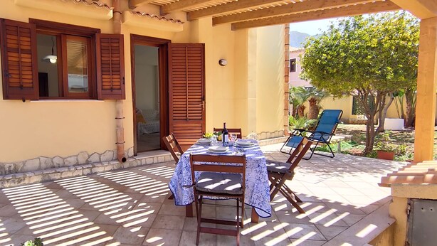 A covered patio with a table set for dining, wooden shutters, and a view of the surrounding area. - Arimondi cottage with patio and garden (San Vito Lo Capo)
