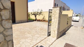 A stone-paved entrance with a gate, a beige building, and a white van parked on the street. - Arimondi cottage with patio and garden (San Vito Lo Capo)