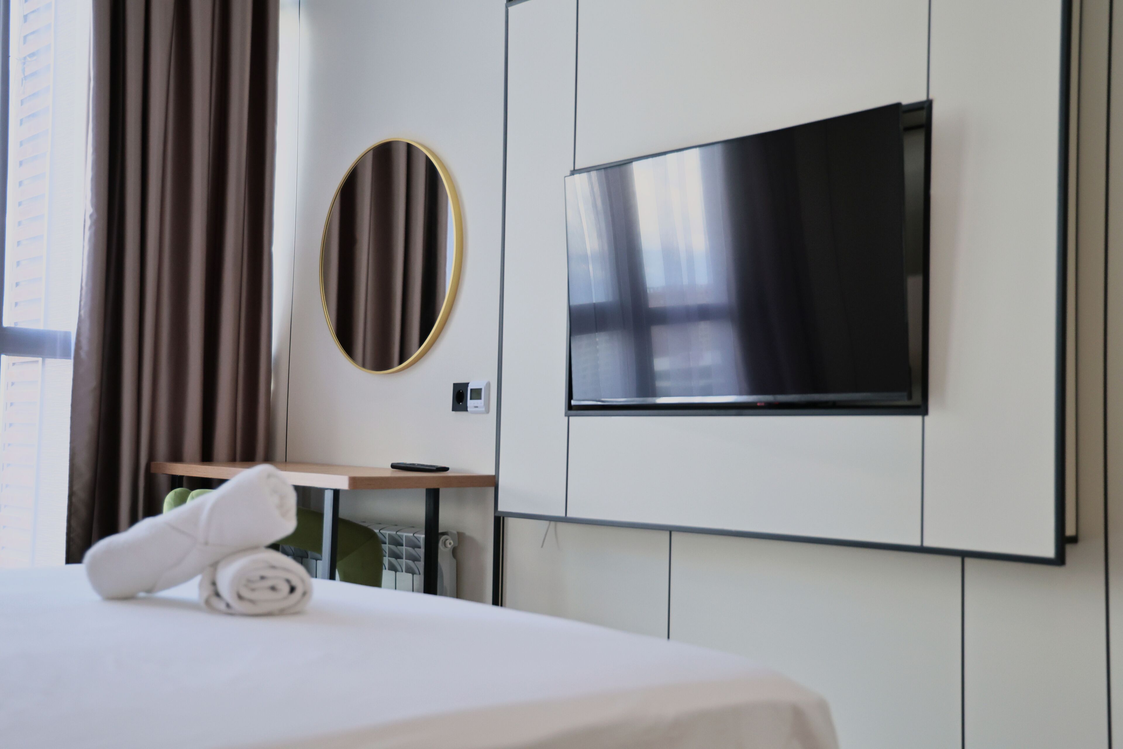 A hotel room with a bed, a TV mounted on the wall, a desk with a chair, and a window with curtains.