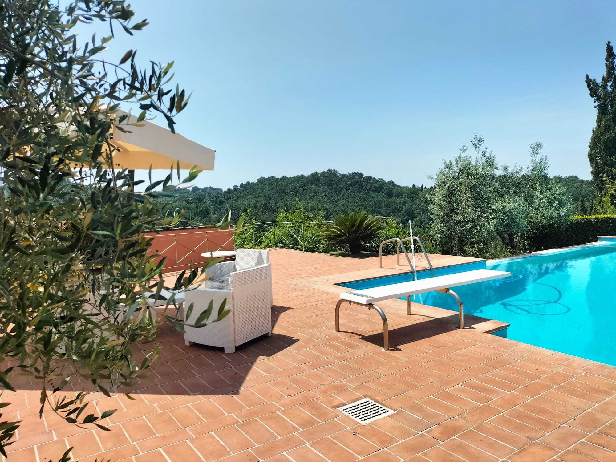 A swimming pool with a white lounge chair and a small table, surrounded by a tiled patio and greenery.