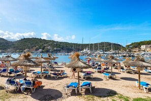 Une plage avec des chaises longues et des parasols en chaume, une marina en arrière-plan et un paysage vallonné.