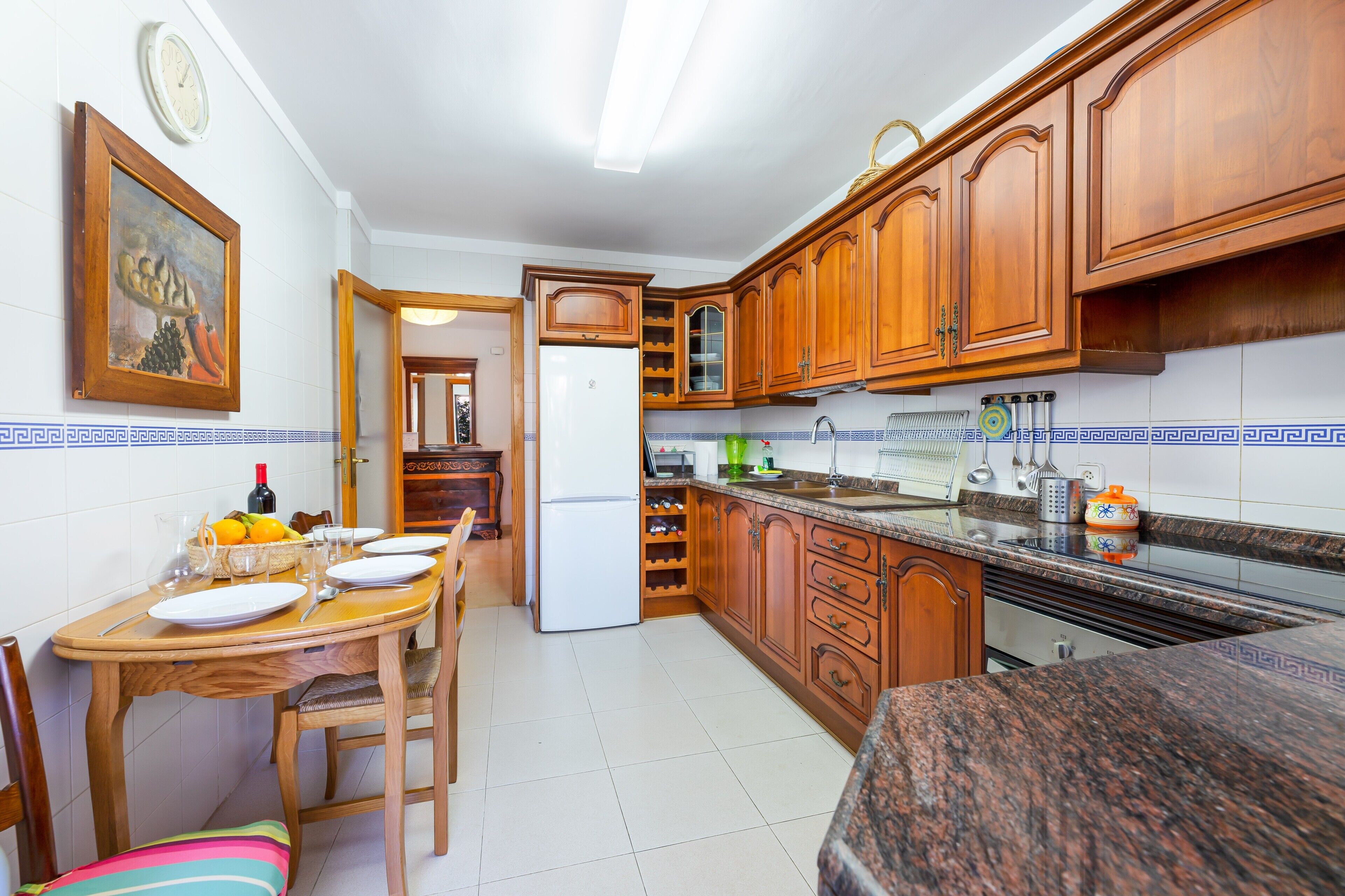 Una cocina con armarios de madera, una mesa de comedor preparada para cuatro comensales, un refrigerador y un reloj en la pared.