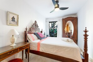 Une chambre à coucher comprenant un lit, une commode, une lampe, un ventilateur de plafond et une fenêtre avec des rideaux.