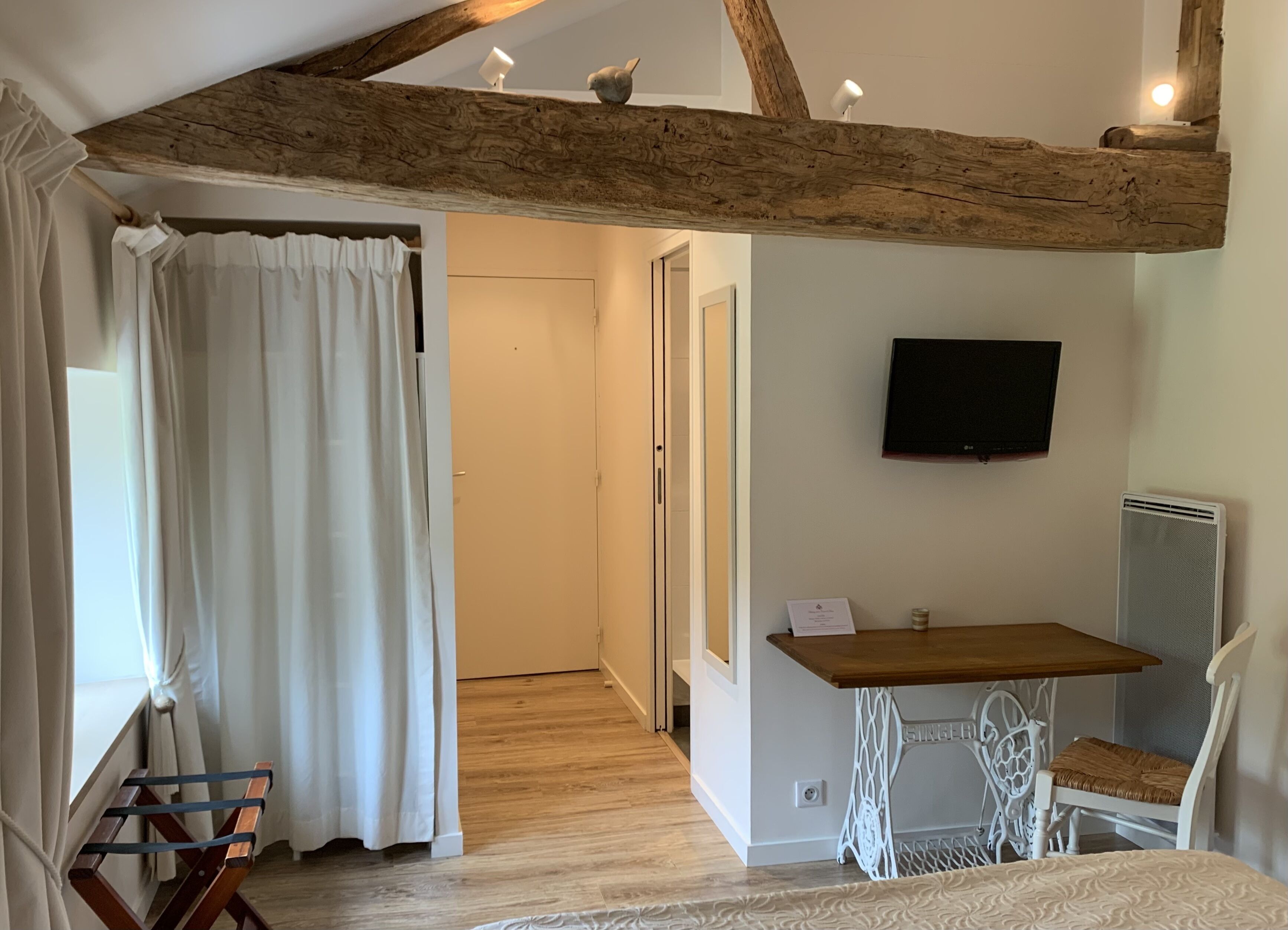 A room with a wooden ceiling beam, a television mounted on the wall, a table with a chair, and a curtain partially drawn.
