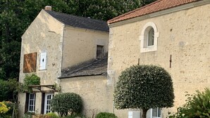 Un bâtiment rustique en pierre, avec un toit de tuiles, une petite fenêtre en arc de cercle et un arbuste taillé avec soin devant.