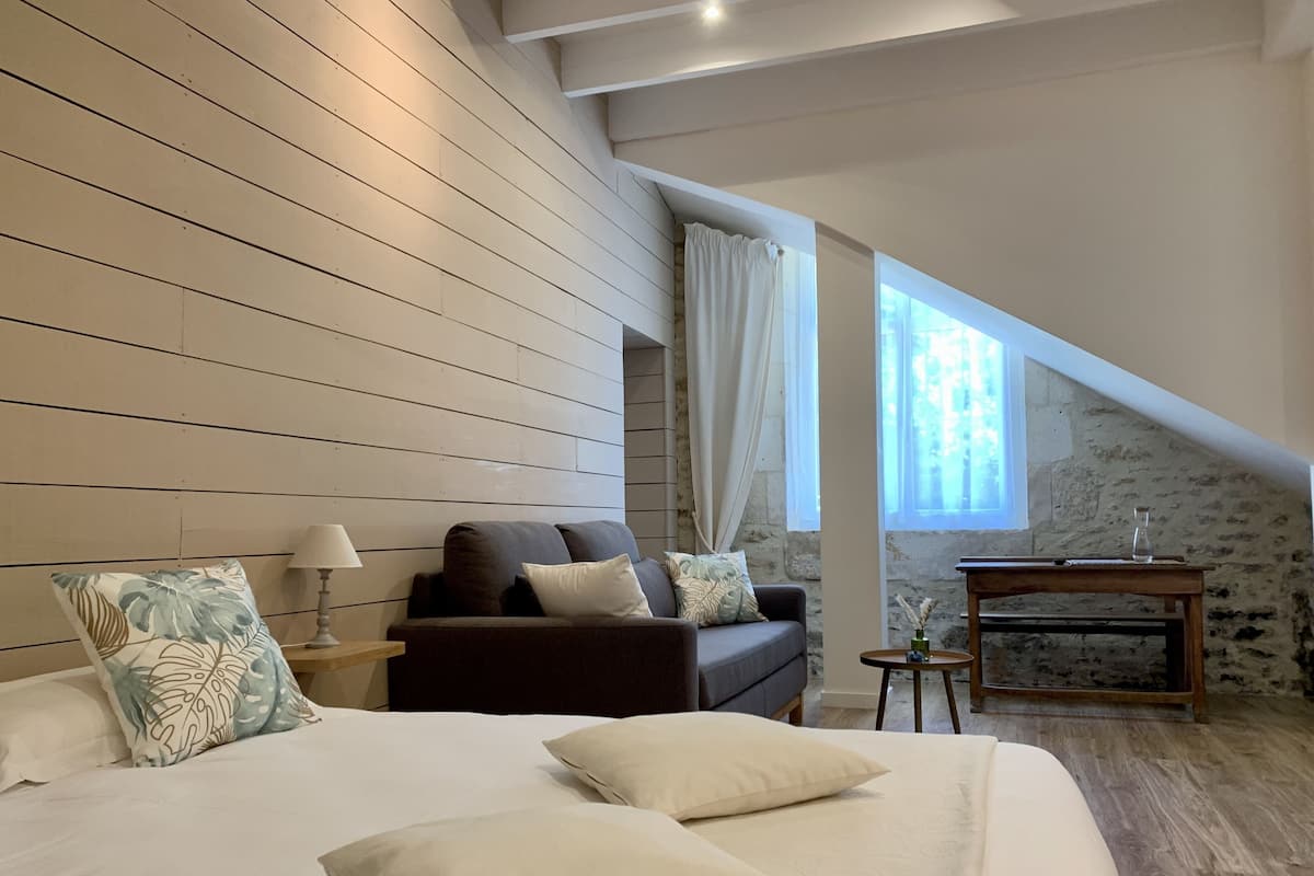 A bedroom with a large bed, a dark sofa, a small table, and a stone wall.