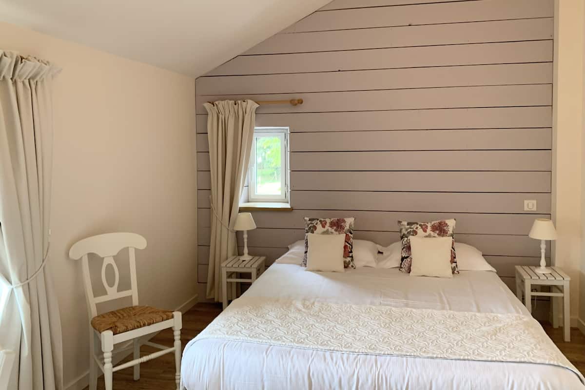 A bedroom with a large bed, two nightstands, a chair, a window with curtains, and a sloped ceiling.