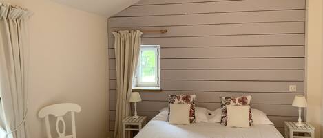 A bedroom with a large bed, two nightstands, a chair, a window with curtains, and a sloped ceiling.