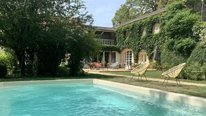 Une piscine à l’eau claire, entourée de verdure et bordée d’un bâtiment au style architectural traditionnel.
