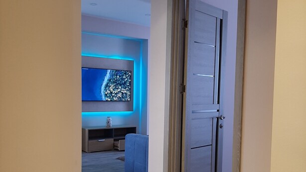 A hallway with a wooden floor, a door, and a room with a TV and blue lighting. - Orexus Tower (Tbilisi)