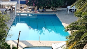 Una piscina rectangular con agua azul cristalina, rodeada por una zona pavimentada con asientos exteriores y una zona cubierta con un letrero que dice 'Welcome'.