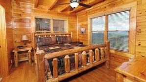 A log cabin bedroom with a wooden bed, a nightstand, a lamp, and a window with blinds. - Scenic Sunrise - Mountain Views + Community Pool Access + Outdoor Hot Tub (Sevierville)