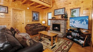 A cozy log cabin interior with a fireplace, leather sofa, and a television. - Scenic Sunrise - Mountain Views + Community Pool Access + Outdoor Hot Tub (Sevierville)