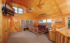 A log cabin bedroom with a bed, a ceiling fan, and a TV mounted on the wall.