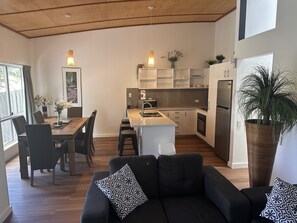 Una cocina moderna con armarios blancos, una isla central y un horno empotrado. Hay un área de comedor con una mesa y sillas de madera, y un sofá negro con cojines estampados.