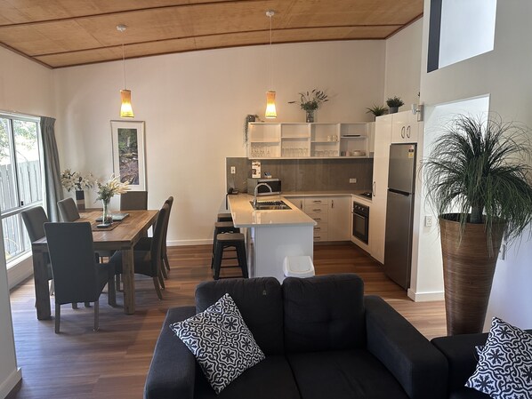 A modern kitchen with white cabinets, a central island, and a built-in oven. There is a dining area with a wooden table and chairs, and a black sofa with patterned cushions. - Sun-Set Satinay at Kingfisher Bay Resort (Kingfisher Bay)