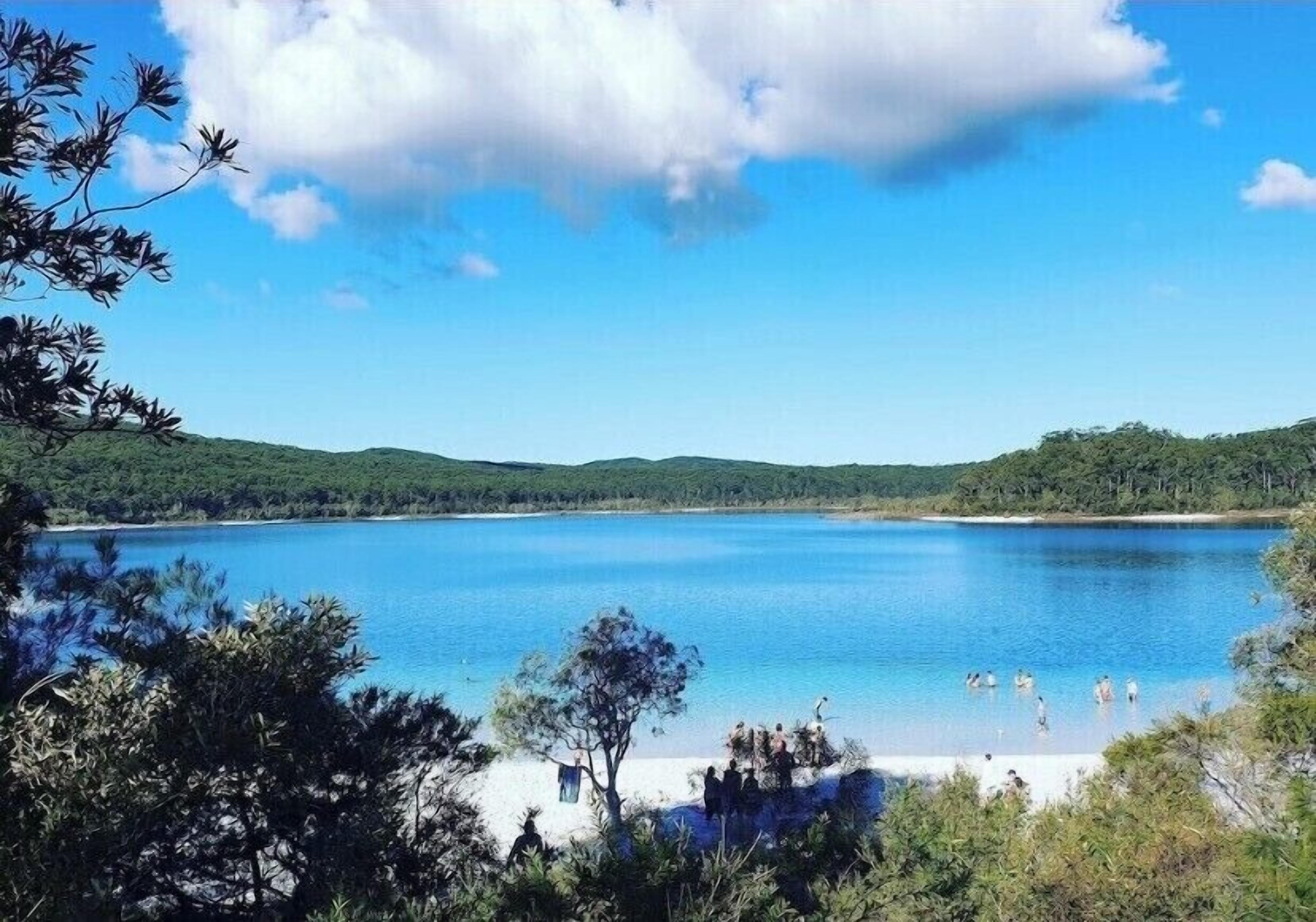 Un lac entouré de collines et d’arbres, où l’on peut voir des gens se baigner et se détendre sur la rive.
