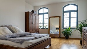 Une chambre à coucher avec un lit en bois, une armoire, un miroir et deux fenêtres surmontées d’arcs.