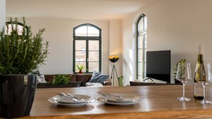 Un coin repas avec une table en bois dressée pour deux personnes, une plante en pot et un téléviseur en arrière-plan.