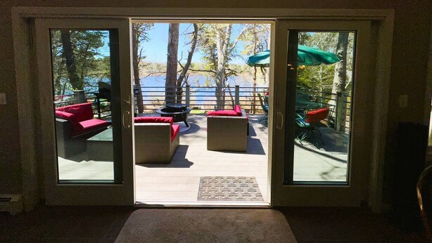 Une porte-fenêtre coulissante donne sur un patio extérieur avec vue sur un lac, agrémenté de meubles d’extérieur rouges et beiges, ainsi que d’un foyer.