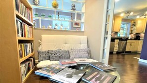 Un salon avec un canapé, une table basse sur laquelle sont posés des magazines, une bibliothèque et, au fond, une cuisine.