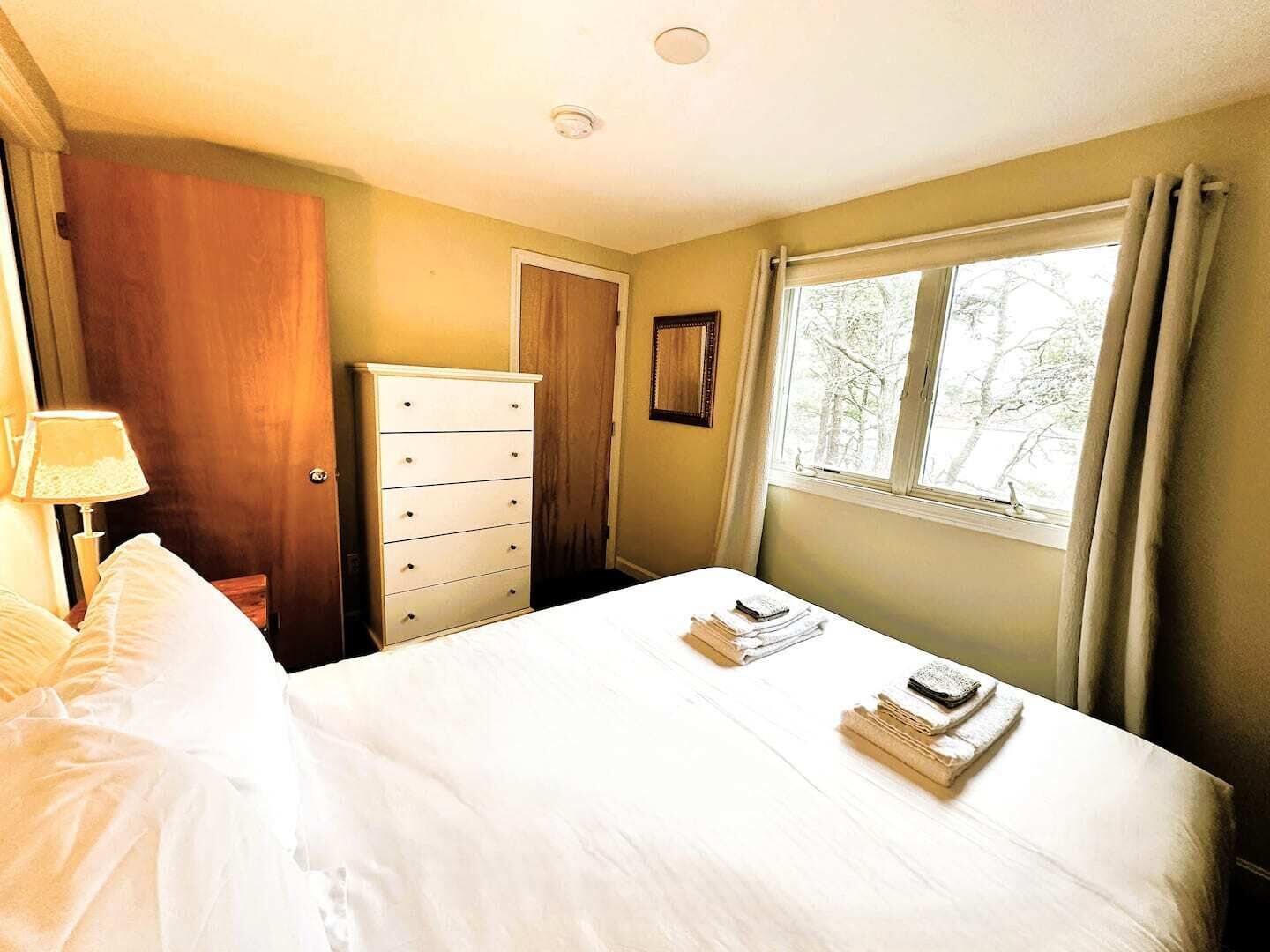 Un dormitorio con cama, cómoda, ventana con cortinas y lámpara.