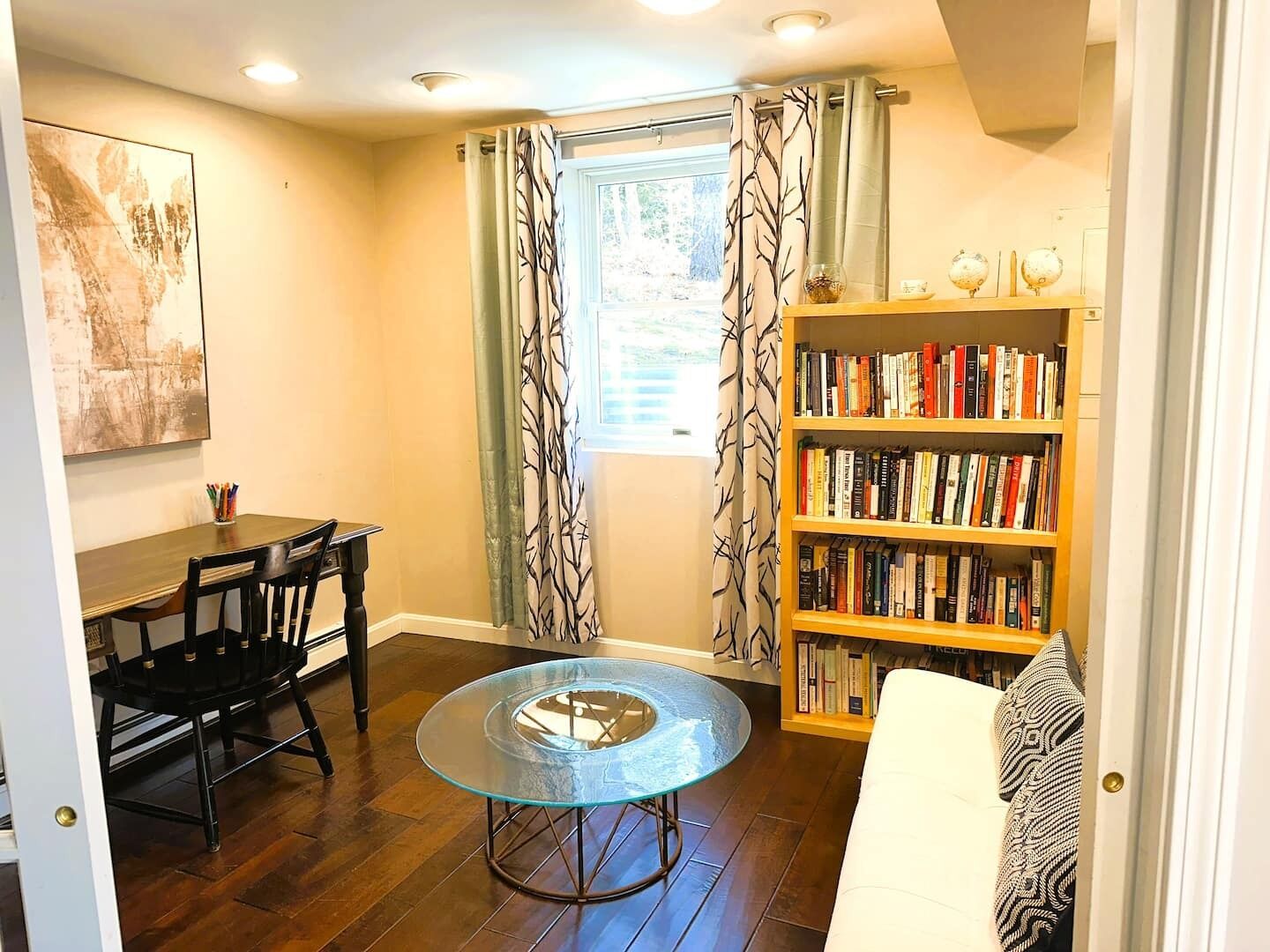 A cozy living room with a round glass table, a bookshelf, a dining area with chairs, and a window with curtains.