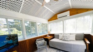 Een kamer met een bed, een televisie, een kleintje tafeltje en een stoel. Er zijn grote ramen met zonwering, een plafond met een geperforeerd patroon en een houten vloer.