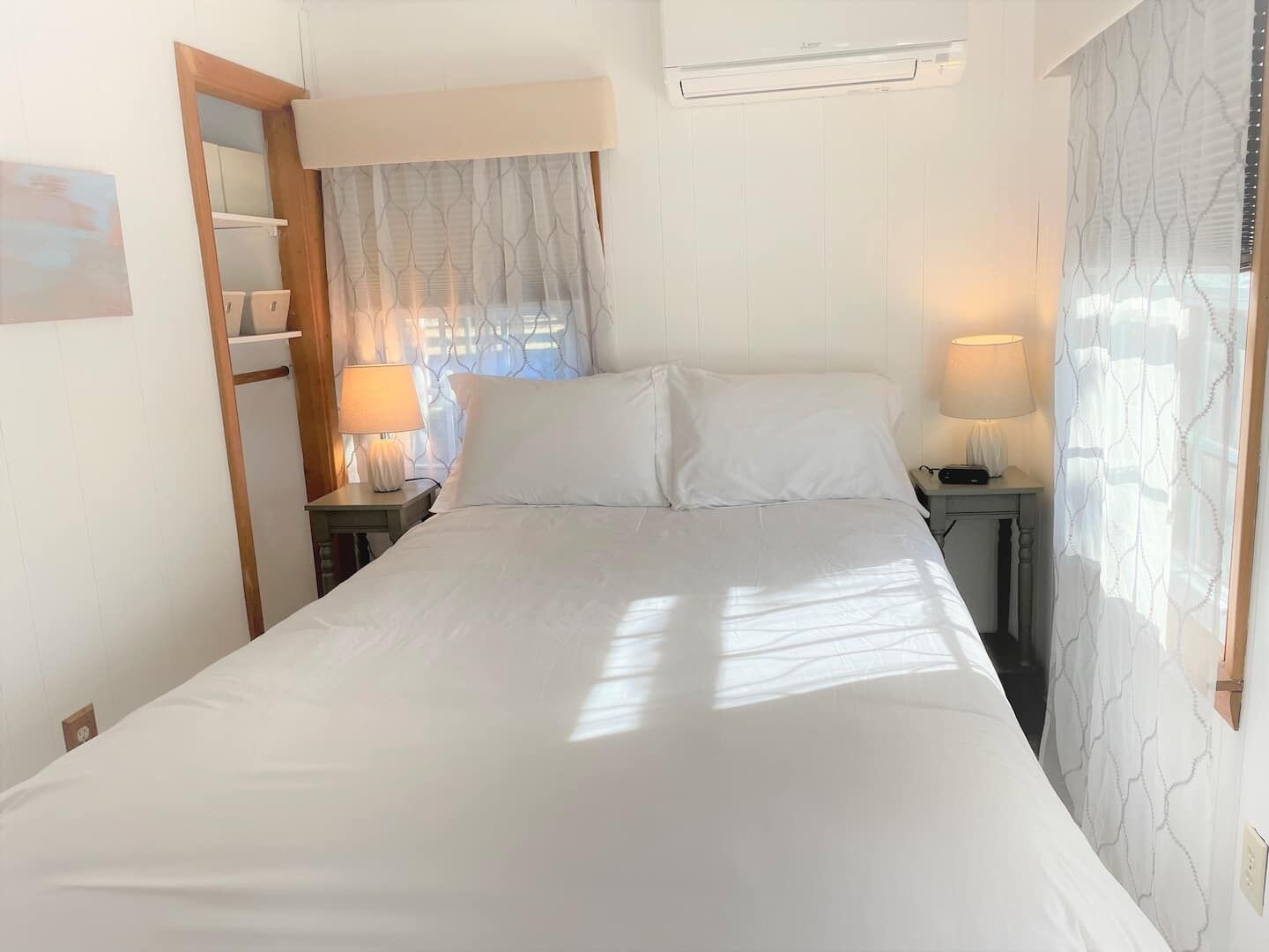 Quarto de hotel com uma cama, duas mesas de cabeceira com luminárias, uma janela com cortinas translúcidas e um ar condicionado na parede.