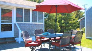 Une terrasse couverte avec une table et des chaises, un parasol rouge et une vue sur une maison dotée d’un porche grillagé.