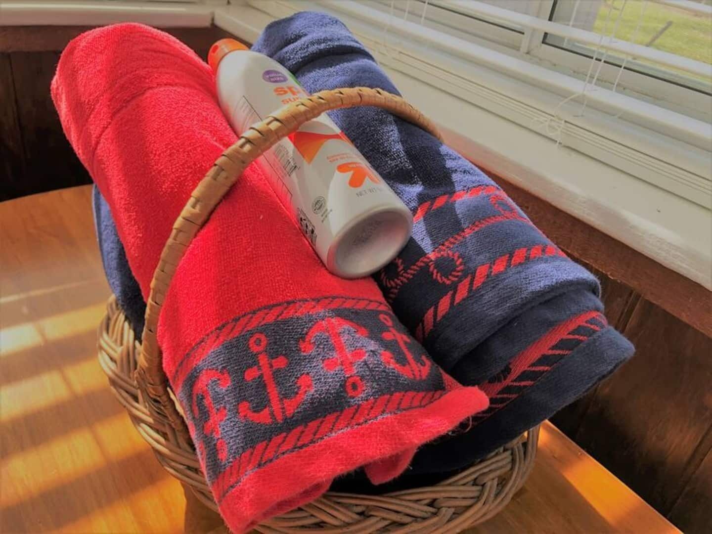Un panier contenant des serviettes enroulées et une bouteille de lotion, posé sur une table en bois près d’une fenêtre.