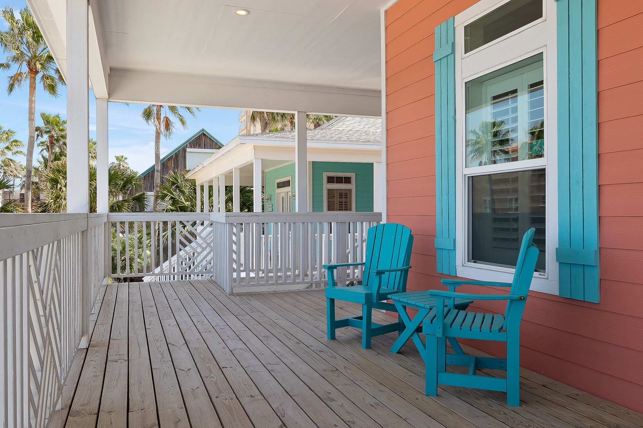 Una terraza de madera con dos sillas Adirondack turquesas y una mesa, con vistas a una zona residencial con casas y palmeras.