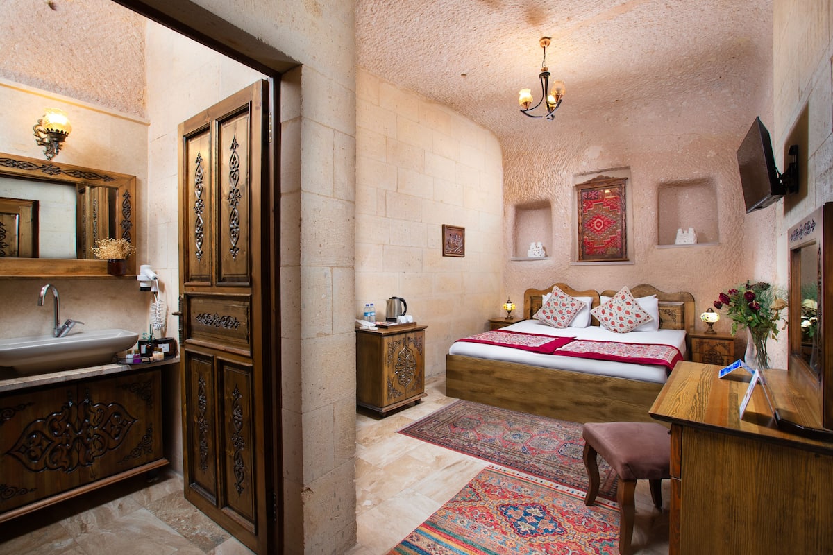 A bedroom with a wooden bed, a nightstand, a desk, a chair, a bathroom with a sink and mirror, and a decorative wall.