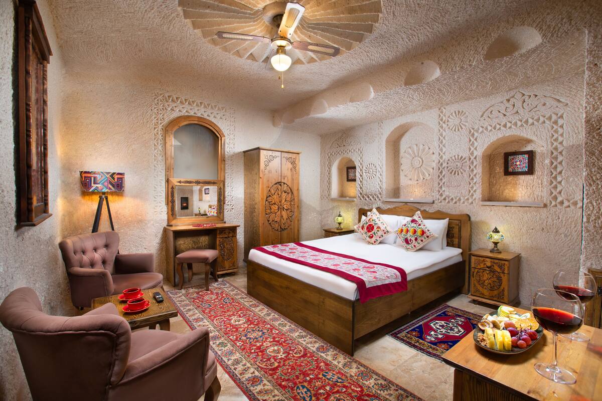 A bedroom with a large bed, a wooden wardrobe, a desk, a chair, and a dining table with fruit and wine.