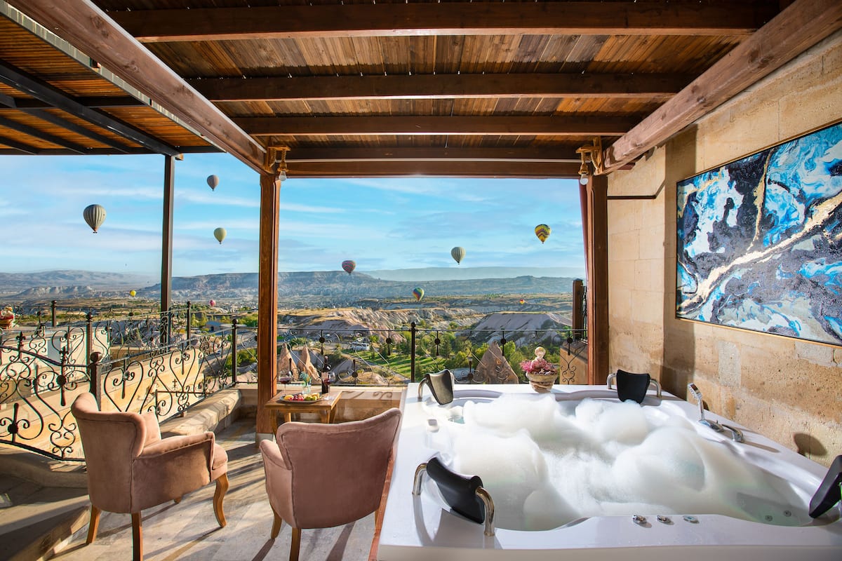A room with a hot tub, two chairs, and a view of hot air balloons over a valley.