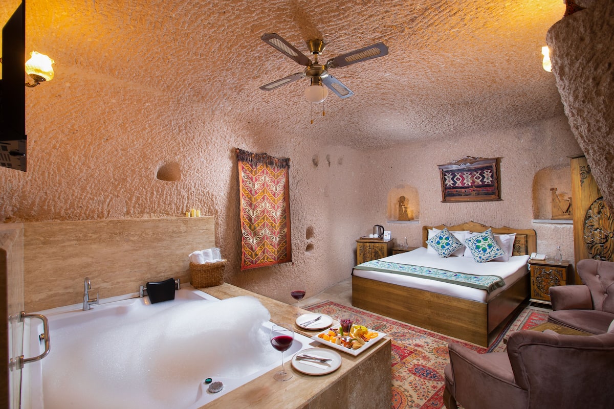 A room with a bathtub, a bed, a ceiling fan, and a small table with food and drinks.