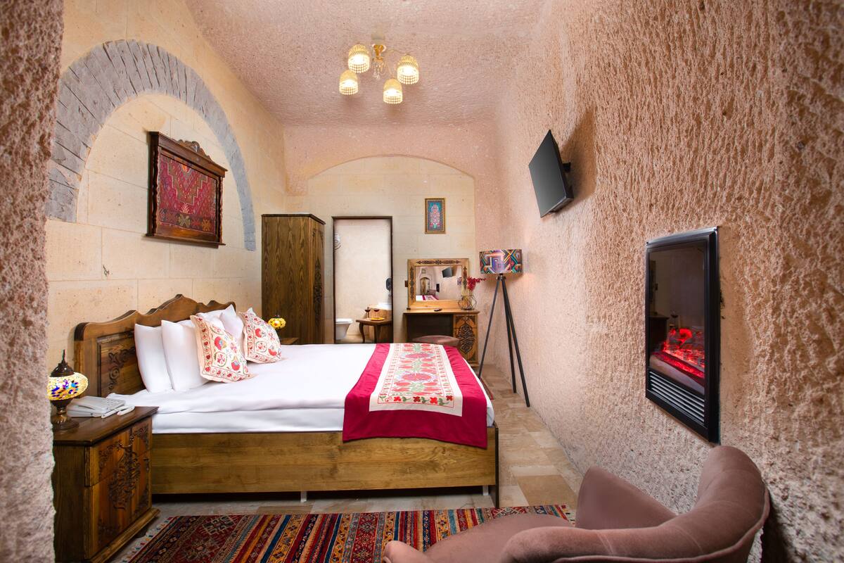A bedroom with a wooden bed, a red and white bedspread, a small table with a lamp, a chair, and a fireplace.