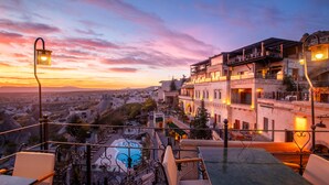 A terrace with a view of a cityscape at sunset, featuring a pool, outdoor seating, and a multi-story building with balconies. - Simera In Cappadocia - Special Class (Nevsehir)