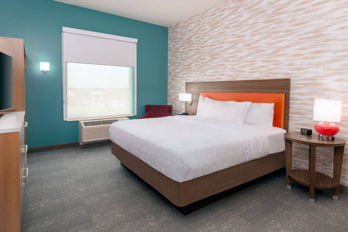 A hotel room with a large bed, a window with blinds, a wall-mounted TV, a bedside table with a lamp, and a red accent wall.