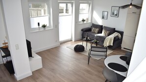 A modern living room with a grey sofa, a small round table, and a white coffee table. There are two windows with blinds, a white cabinet, and a black floor lamp. - Beautiful vacation apartment near Pietzmoor (Schneverdingen)