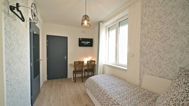 Ein Schlafzimmer mit einem Bett, einem Schreibtisch mit zwei Stühlen, einem Fernseher und einem Fenster mit Vorhängen.