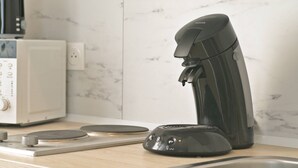 Eine Kaffeemaschine steht auf einer Küchenarbeitsplatte, neben einem weißen Gerät und einer Marmor-Spritzschutzwand.