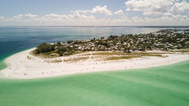 Une vue aérienne d’une ville côtière avec une plage de sable, des eaux turquoise et limpides, et un ensemble dense de bâtiments.