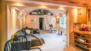Una sala de estar acogedora con un sofá, una silla, una escalera y una zona de cocina con estufa y estanterías.