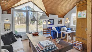 A cozy cabin interior with a wooden ceiling, a dining area, a sofa, and a table with books.