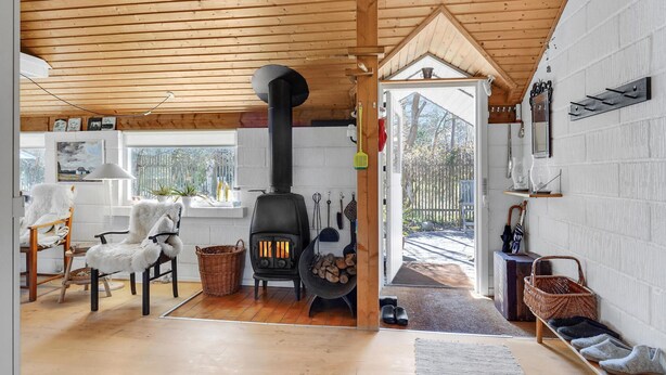 A cozy cabin interior with a wood-burning stove, rustic furniture, and a view through a large door to the outside.