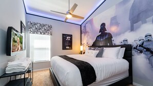 Une chambre à coucher avec un lit, un ventilateur de plafond, des œuvres d’art murales et une fenêtre avec des stores.