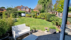 Een terras met een witte wickerbank, een metalen zitbank en een kleine tafel. Er staan potplanten en een verzorgd gazon op de achtergrond.