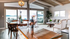 A dining area with a wooden table, chairs, and a wine bottle. There are large windows and a view of the outdoors. - Lakefront Family Getaway | Dock, Hot Tub, Kings (Lakeport)