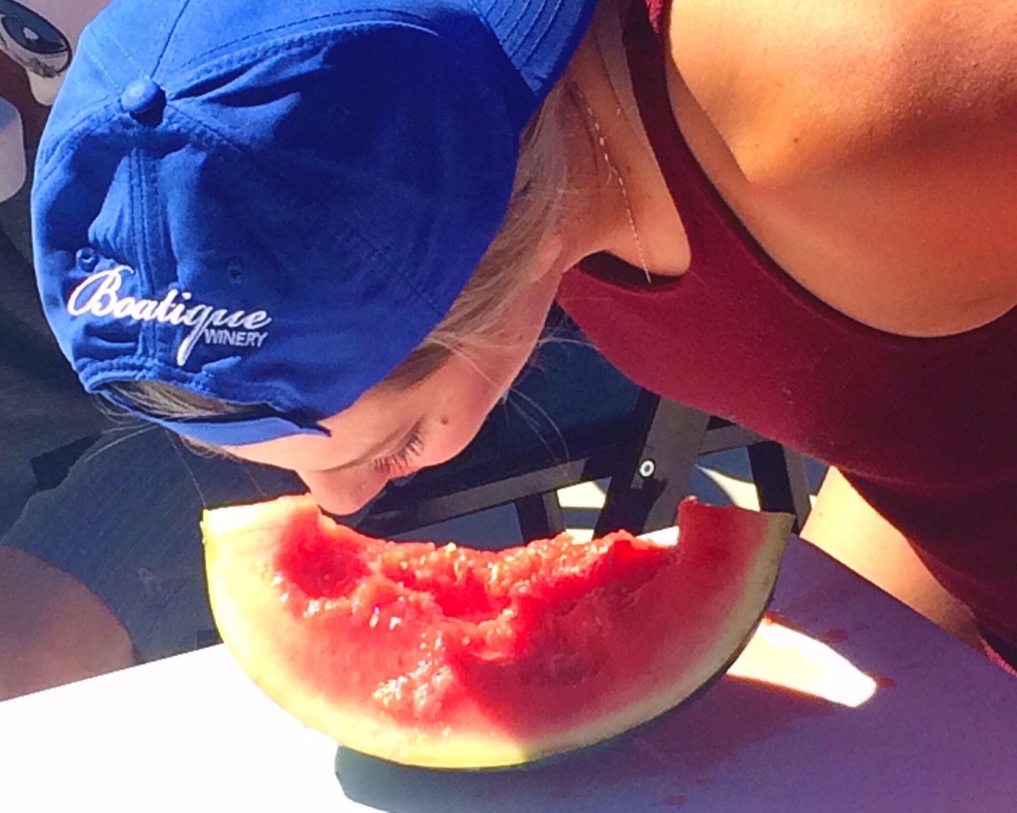 A person wearing a blue cap with a logo, eating a slice of watermelon.
